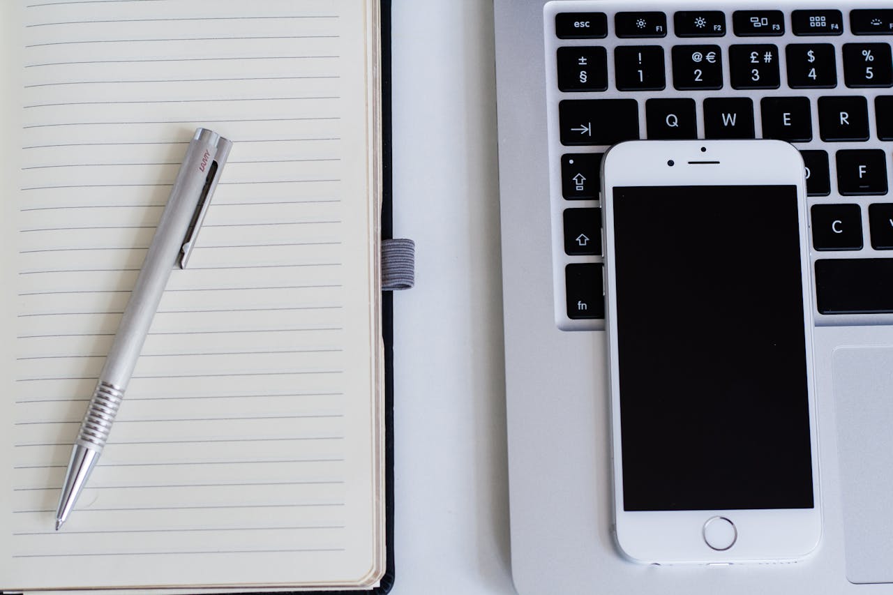 Como fugir da produtividade tóxica? 6 Notebook, celular, caderno e caneta em cima de uma mesa branca. Essa imagen representa as ferramentas e recursos recomendados no artigo.