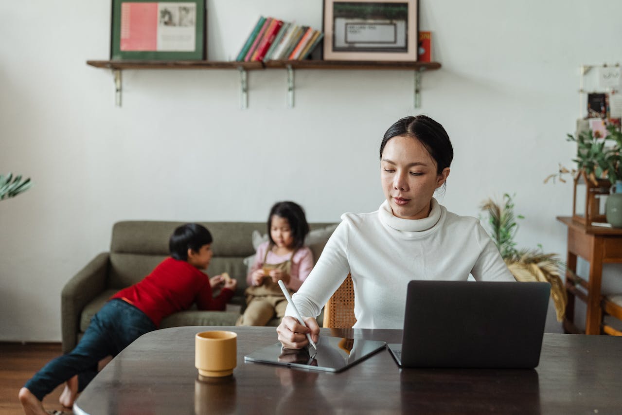 Como manter uma rotina diária com apenas 15 minutos por dia 2 Muler sentada em uma mesa mexendo no tablet e os filhos ao fundo brincando no sofá. A imagem representa o tópico do artigo "Por que manter uma rotina diária, mesmo que breve?"