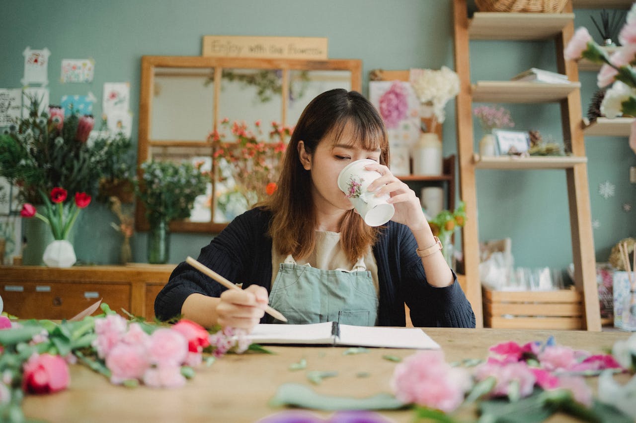 Como manter uma rotina diária com apenas 15 minutos por dia 3 Moça em uma floricultura sentada em uma mesa de madeira, escrevendo em um caderno. Essa imagem representa o tópico do artigo "Ideias de micro-rotinas para se inspirar"